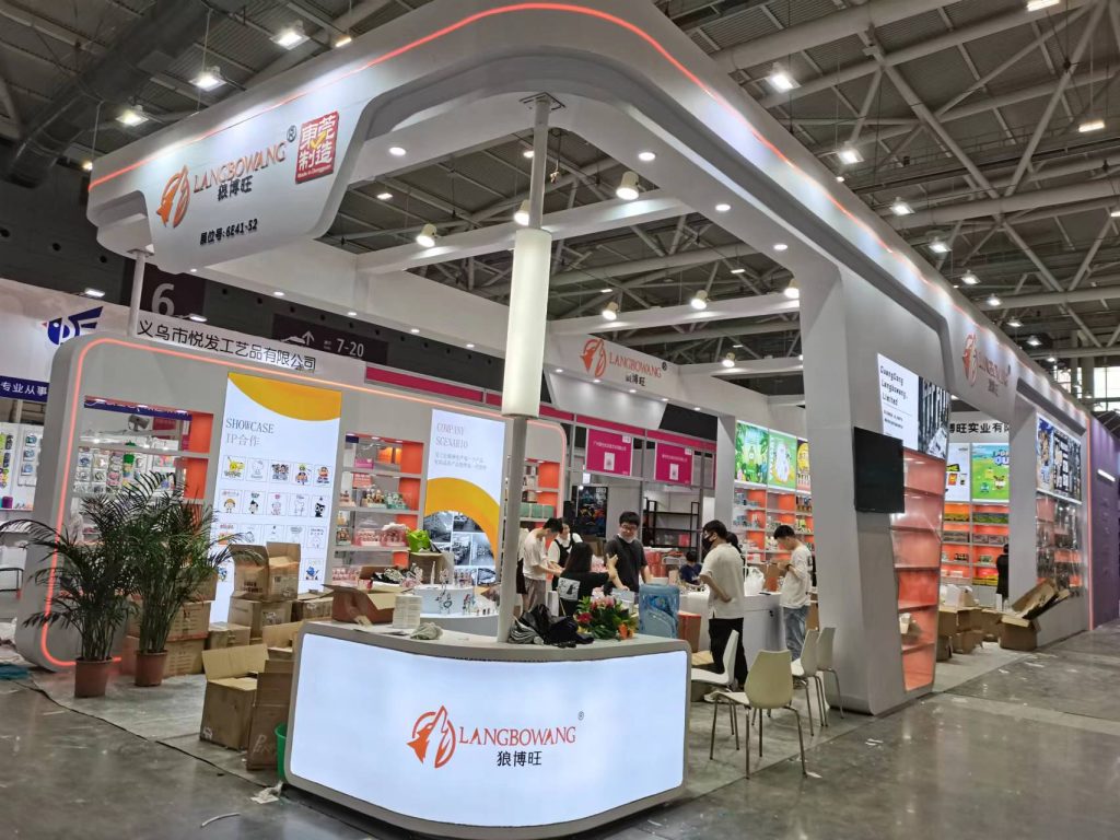 Exhibition booth with a reception desk, illuminated displays, and people setting up and browsing. Construction materials are on the floor.