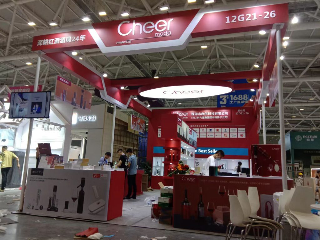 A red and white exhibition booth with display items, workers setting up, and scattered boxes in a large hall.
