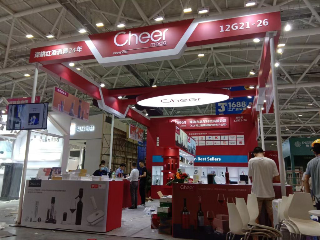 A red and white exhibition booth with workers arranging products and boxes, set in a spacious exhibition hall.