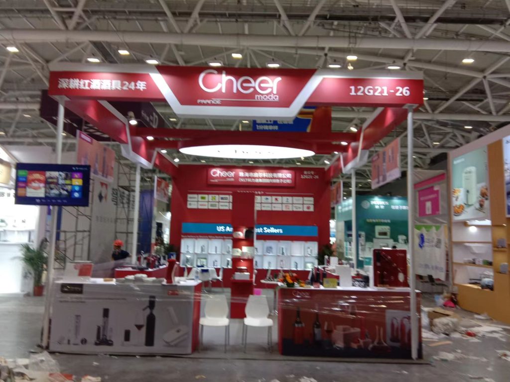 A blurry wide shot of a red and white exhibition booth during setup in a large convention hall.