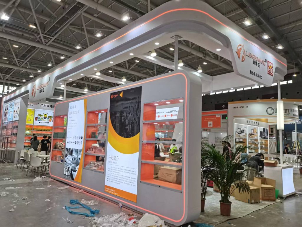 Exhibition booth with orange LED lighting during setup, showing display shelves, promotional posters, and scattered construction materials.