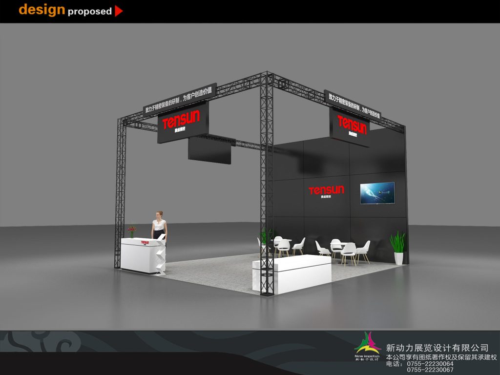 Angled view of an exhibition booth with black walls, a screen, white reception desk with a mannequin, and a seating area under a metal truss frame.
