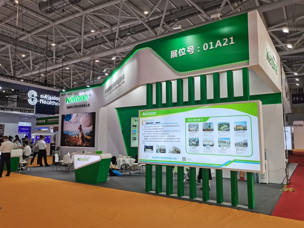 A modern white and green trade show booth with a large display screen and information panels. Attendees are visible in the background.