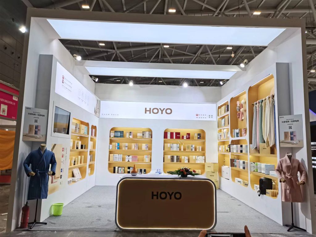 Exhibition booth with white and gold shelves displaying numerous boxed products. Two mannequins wear blue and pink robes.
