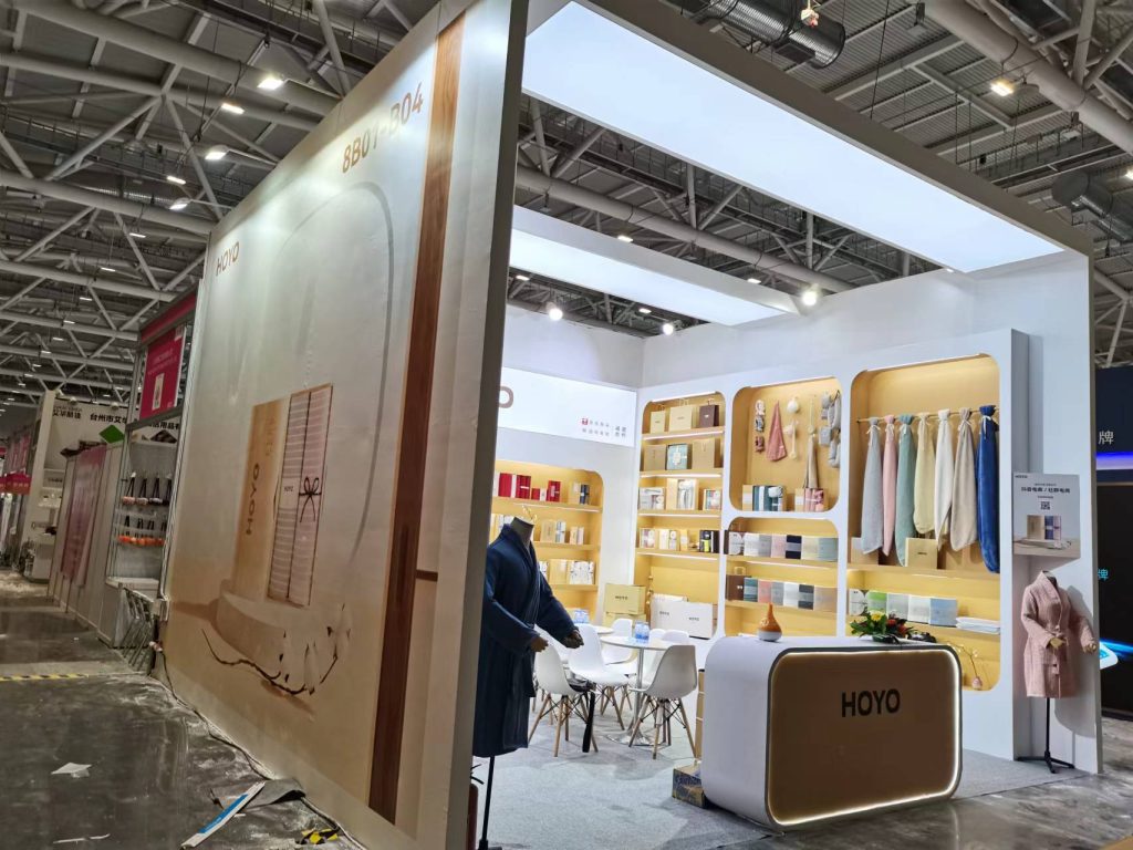 Side view of an exhibition booth with a mannequin in a blue robe, small white tables, and product displays visible inside.