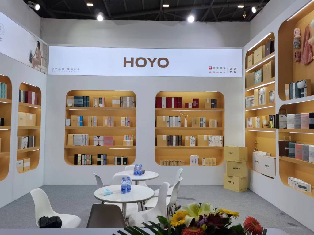 Interior of a white exhibition booth, showcasing multiple shelves of packaged products, small round tables with chairs, and a floral centerpiece.