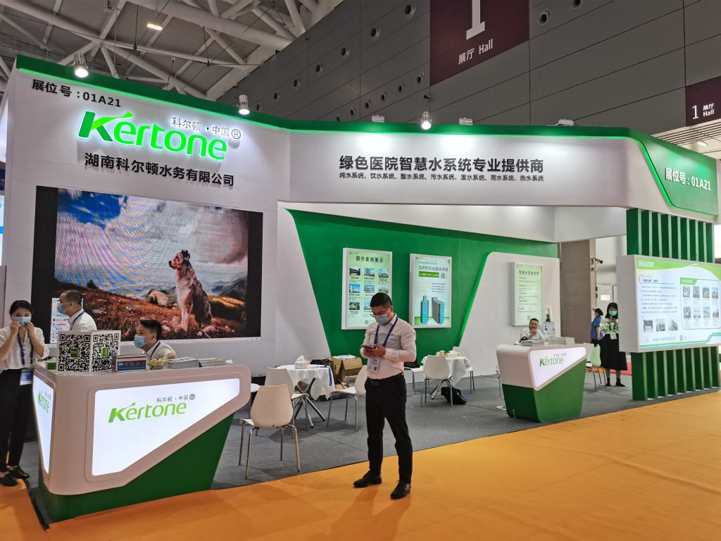 A white and green trade show booth with a large screen displaying a dog. Several attendees are present, one looking at a phone.