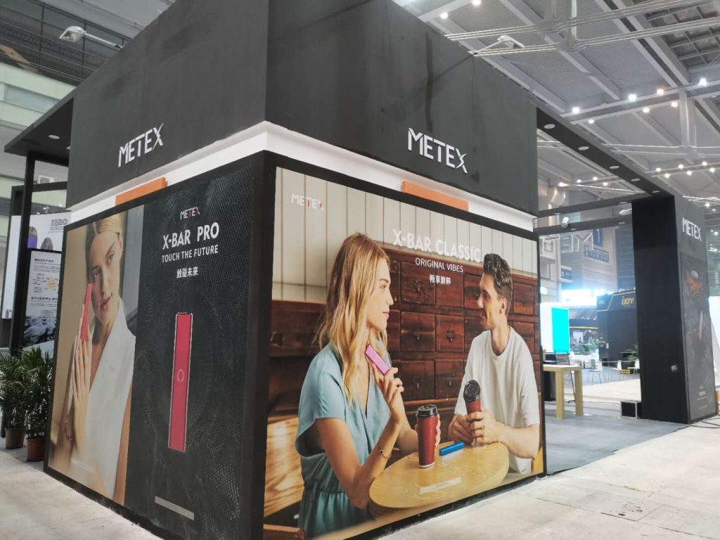A black trade show booth featuring large posters of people using pink handheld devices, set in an exhibition hall.