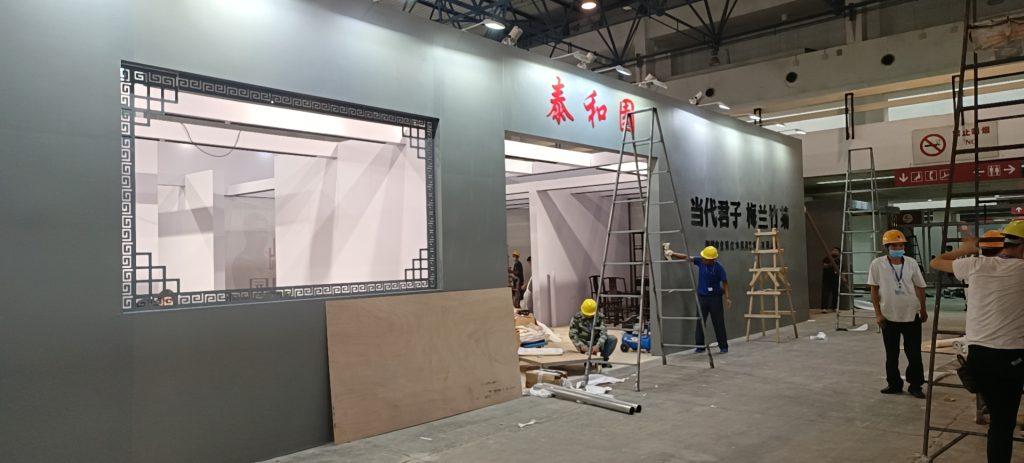 Exhibit booth under construction. Workers on ladders, plywood, and scaffolding surround a wall with a decorative frame and Chinese characters.
