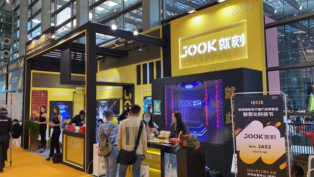 Busy yellow and black trade show booth with glowing displays and attendees interacting.