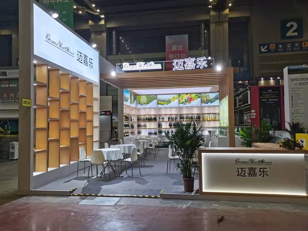 A large, brightly lit exhibition booth inside a hall, featuring seating areas, display shelves filled with bottles, and potted green plants.
