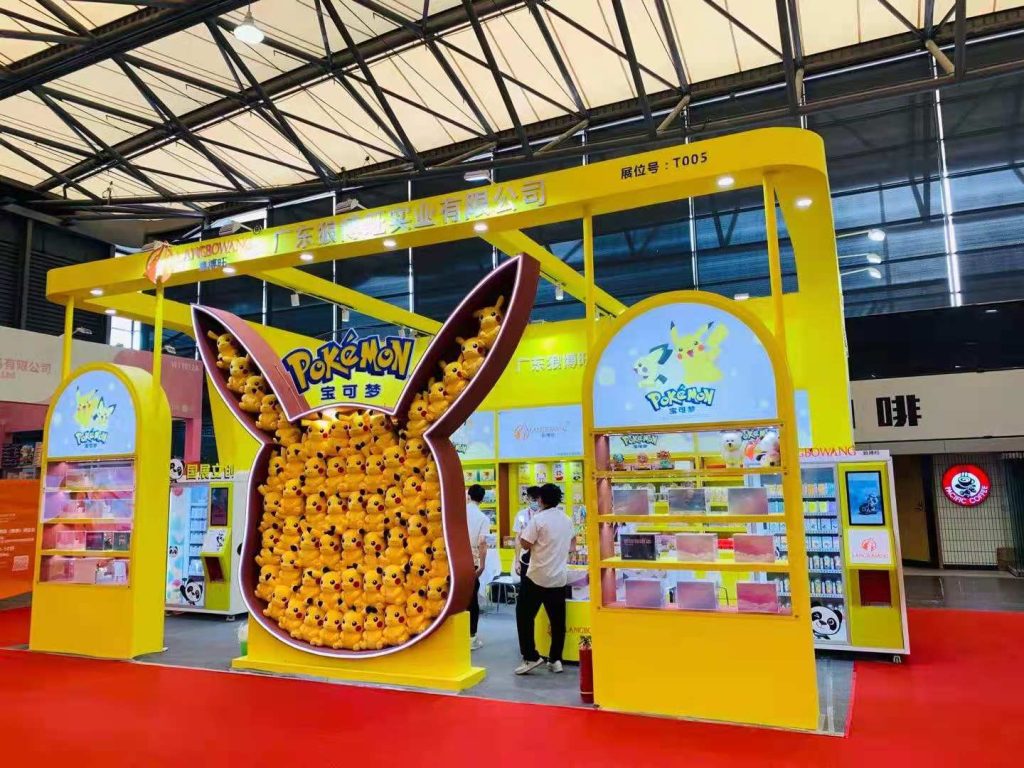 Yellow exhibition booth with a large, ear-shaped display full of yellow plush toys. People browse merchandise at the brightly lit fair.