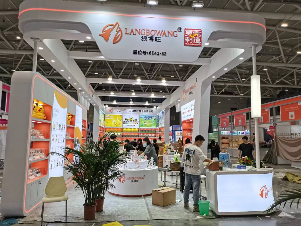 People at a bustling exhibition booth with orange and white displays, products on shelves, a reception desk, and decorative plants.