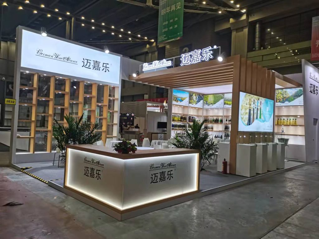 Exhibition booth with a glowing white reception desk, several potted green plants, white tables, and display shelves showcasing various products.
