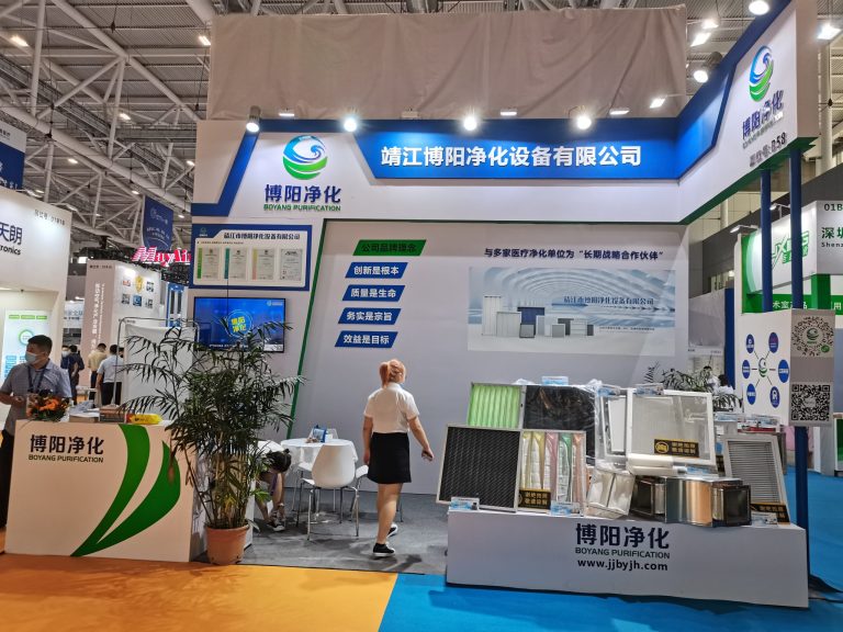 Trade show booth displaying air filters and purification devices. People walk past plants and displays in a busy exhibition hall.