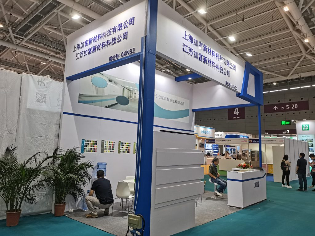 Exhibition booth in a convention hall, with two men viewing product samples near potted plants. Other attendees are visible in the background.