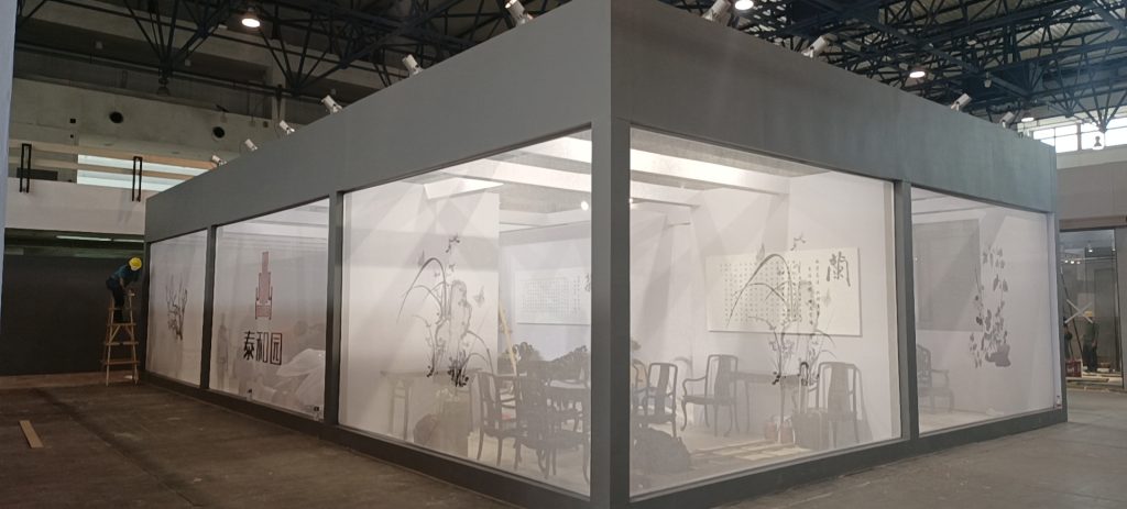 Constructed exhibit booth with large glass walls. Inside, traditional Chinese artwork and wooden furniture are visible.