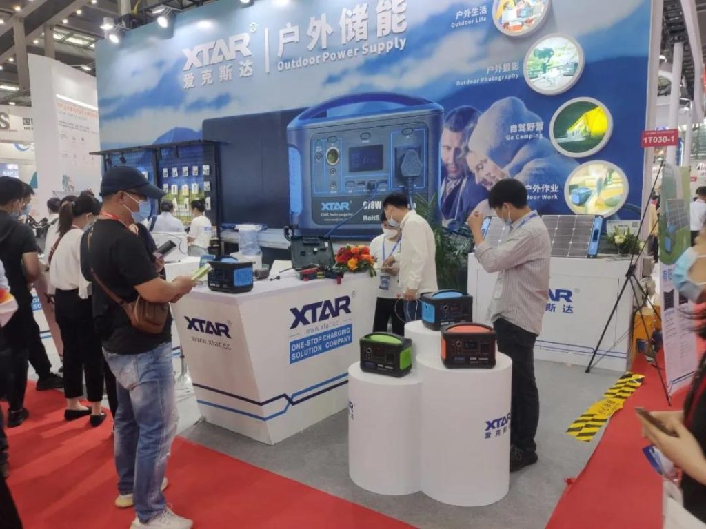 People viewing outdoor power supplies at a lively exhibition booth with a large blue banner and white display pedestals.