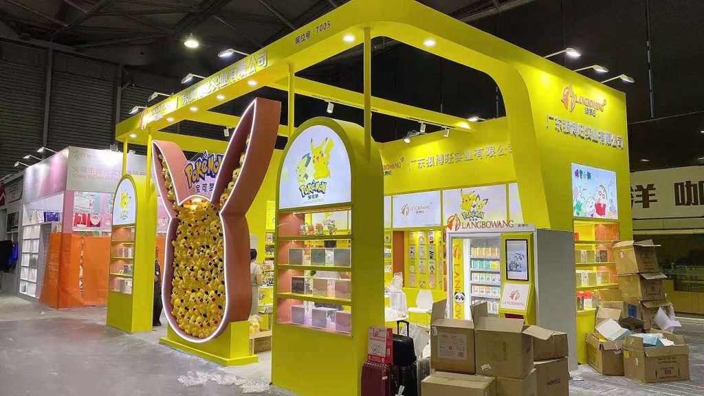 Yellow exhibition booth with an ear-shaped display packed with yellow plush toys. Empty shelves, stacked boxes, and luggage are visible at the trade show.