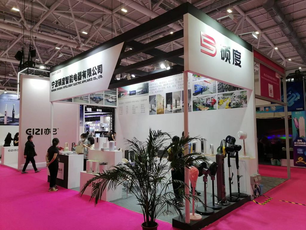 Exhibition booth at a trade show, with attendees, various household appliances on display, and a large potted plant on a pink carpeted floor.