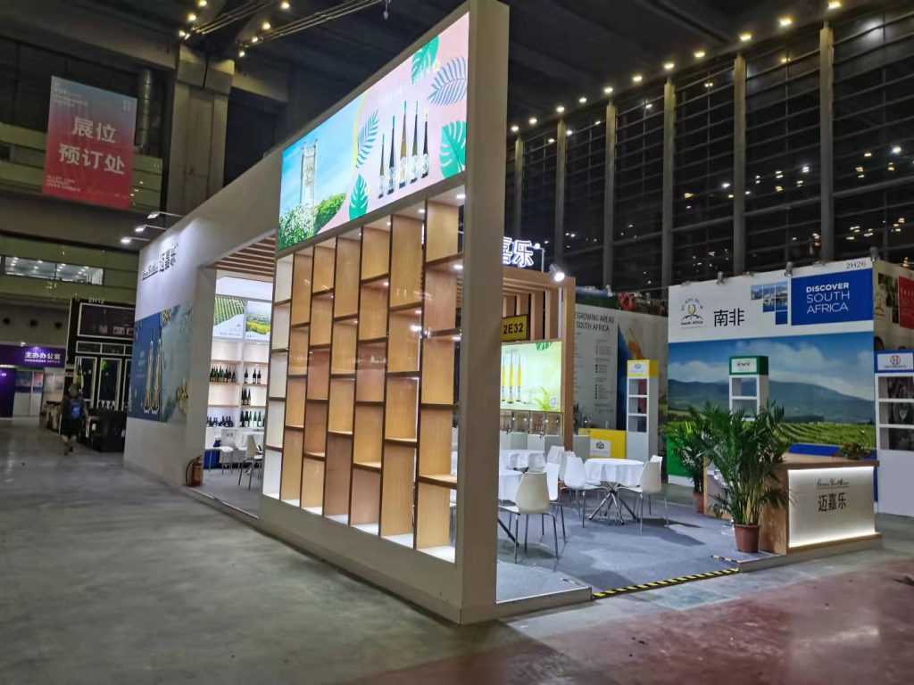Side view of an exhibition booth with a tall wall of square wooden cubbies, large digital screens above, and a seating area with white tables and chairs.