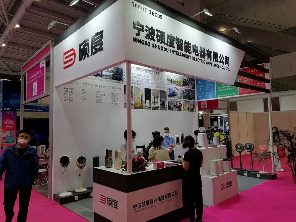 People interact at an exhibition booth counter, surrounded by displayed white and pink household appliances, with a large potted plant in the foreground.