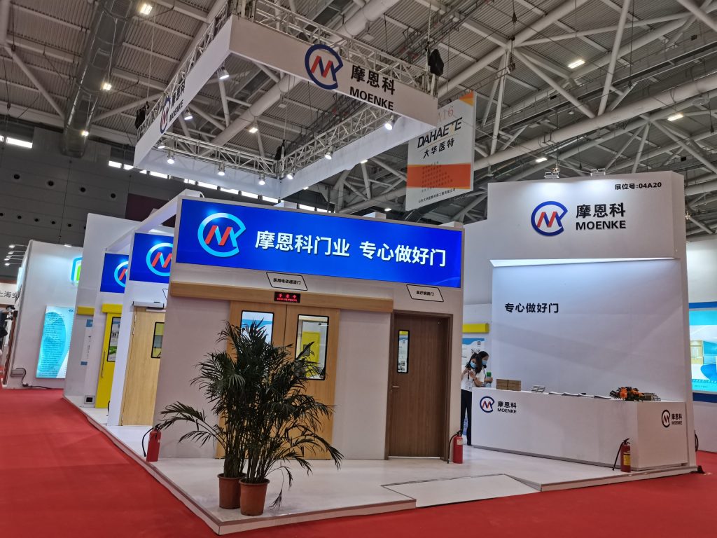 An exhibition booth displaying various door types, including automatic and wood-paneled, with a plant and a reception desk. Red carpet in the foreground.