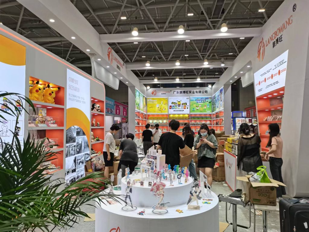 Crowded trade show booth with visitors browsing products on multi-tiered shelves and circular display tables with figures.