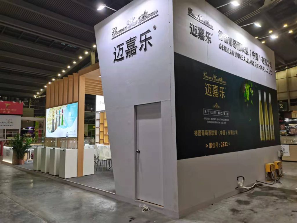 Exterior wall of an exhibition booth with a white closed door, large dark panels displaying text and images of bottles, and visible wires on the floor.