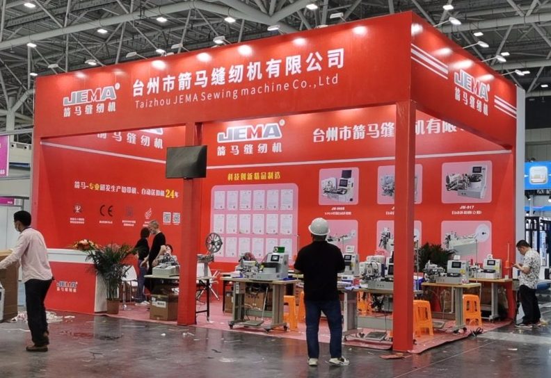 People interact with industrial sewing machines inside a red and white exhibition booth.