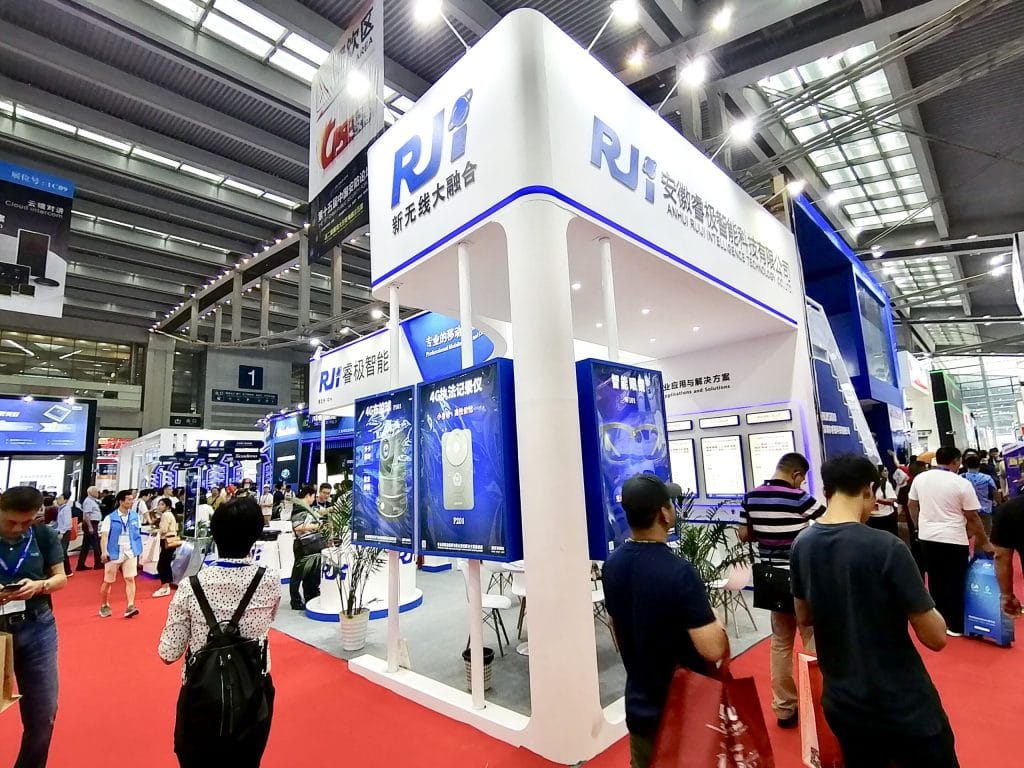 Busy exhibition hall with a prominent white and blue booth. People browse displays on a red carpeted floor under an industrial ceiling.