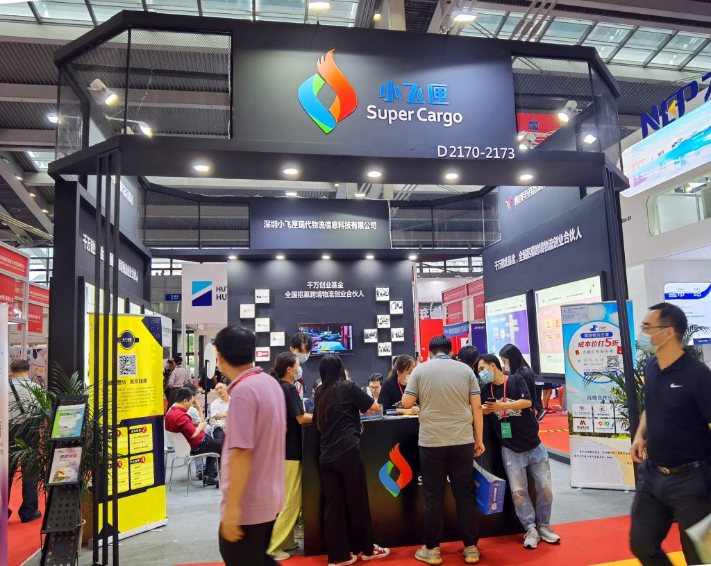 A busy dark trade show booth with an illuminated sign and multiple screens, surrounded by attendees on a red carpet.