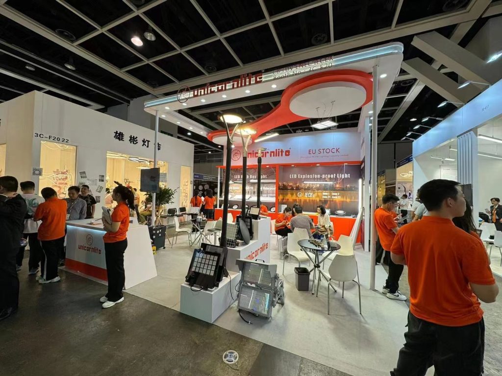 People gather in a lively trade show booth with orange accents, displaying various lighting products, a counter, and seating.