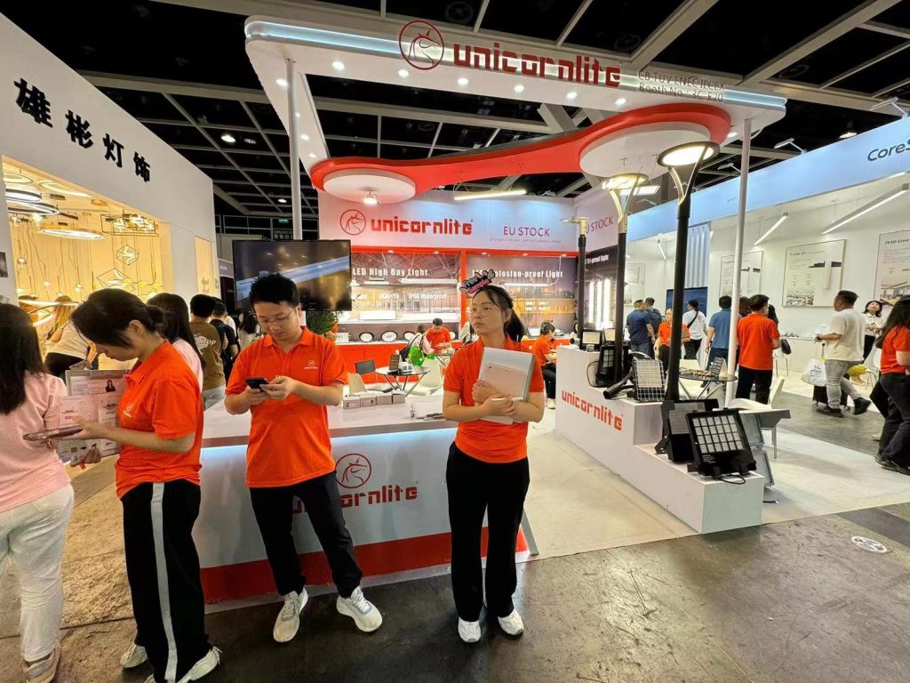 People interact at a busy trade show booth with a white counter and orange branding, showcasing light fixtures.