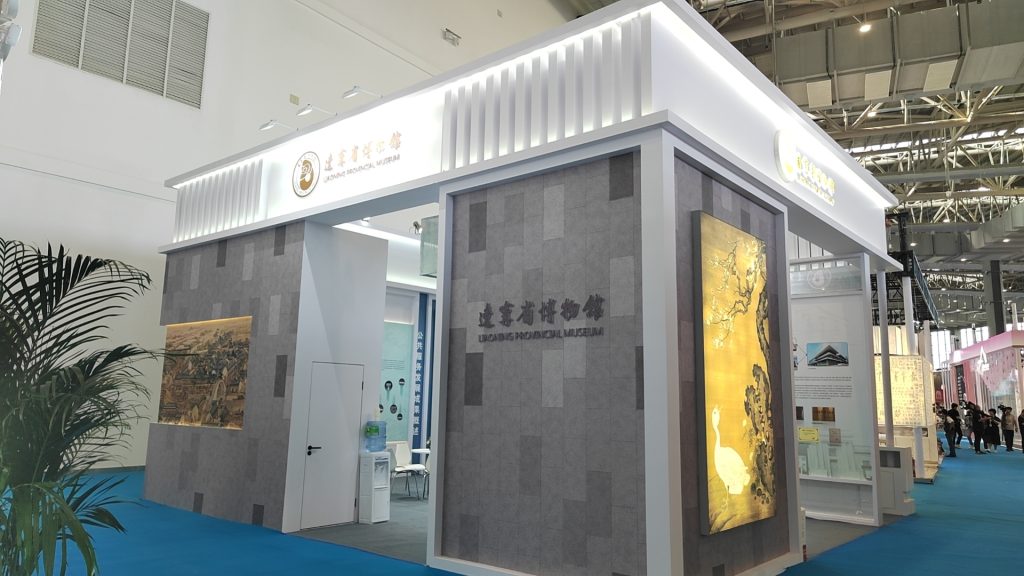 Exhibition booth with gray walls, white trim, and a prominent "PROVINCIAL MUSEUM" sign, displaying golden art panels with a bird and a historical scene.
