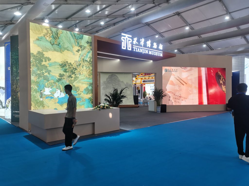 Exhibition hall with a large, elaborate booth featuring a glowing "TIANJIN MUSEUM" sign, traditional art panels, and people walking on a blue carpet.
