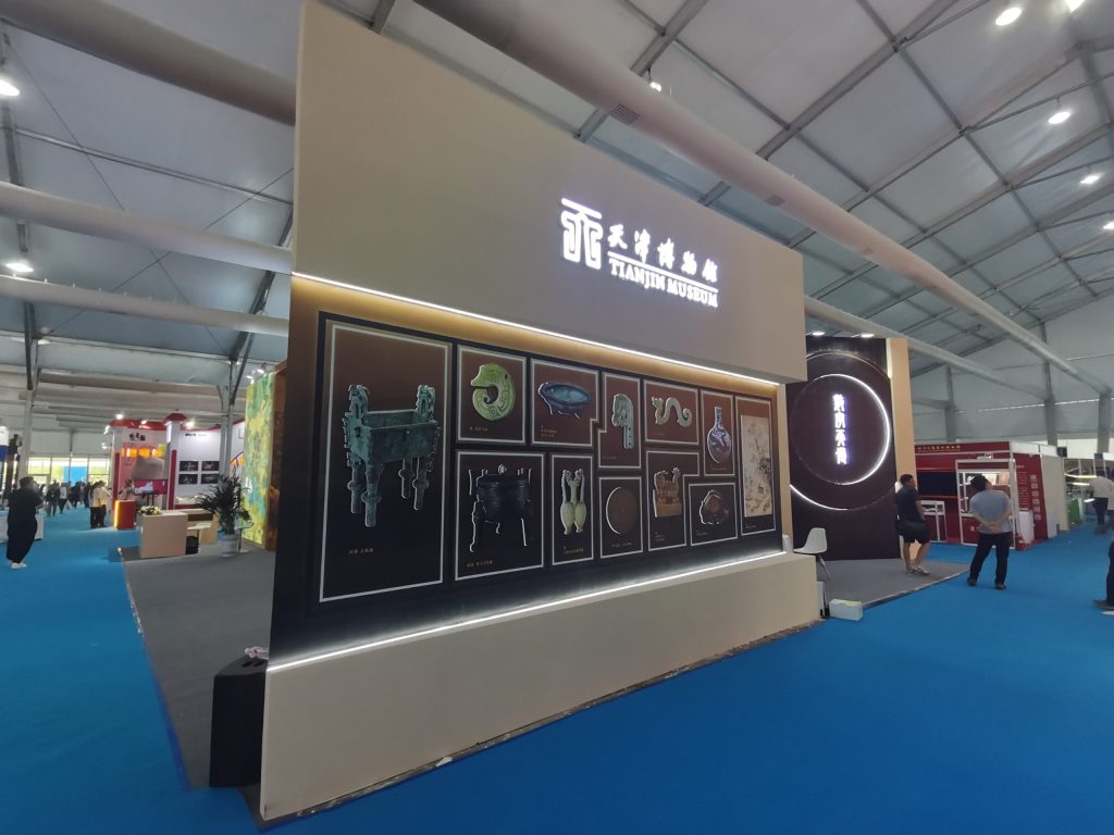 View of the "TIANJIN MUSEUM" booth, highlighting a display wall with framed images of ancient artifacts like bronze vessels and jade carvings.
