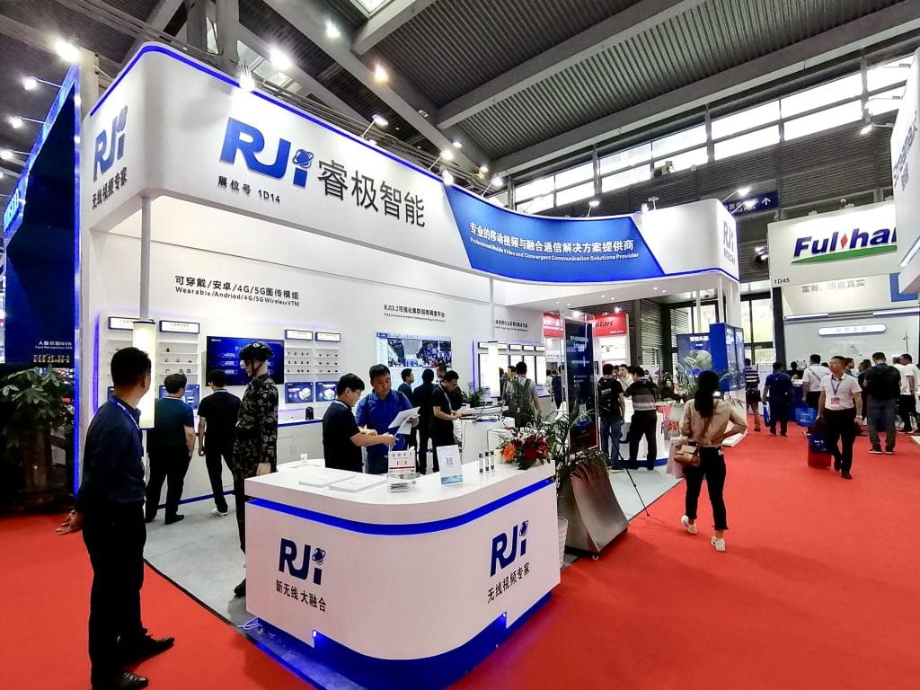 Wide view of a white and blue exhibition booth in a crowded hall. People gather at counters and displays on a red carpet.
