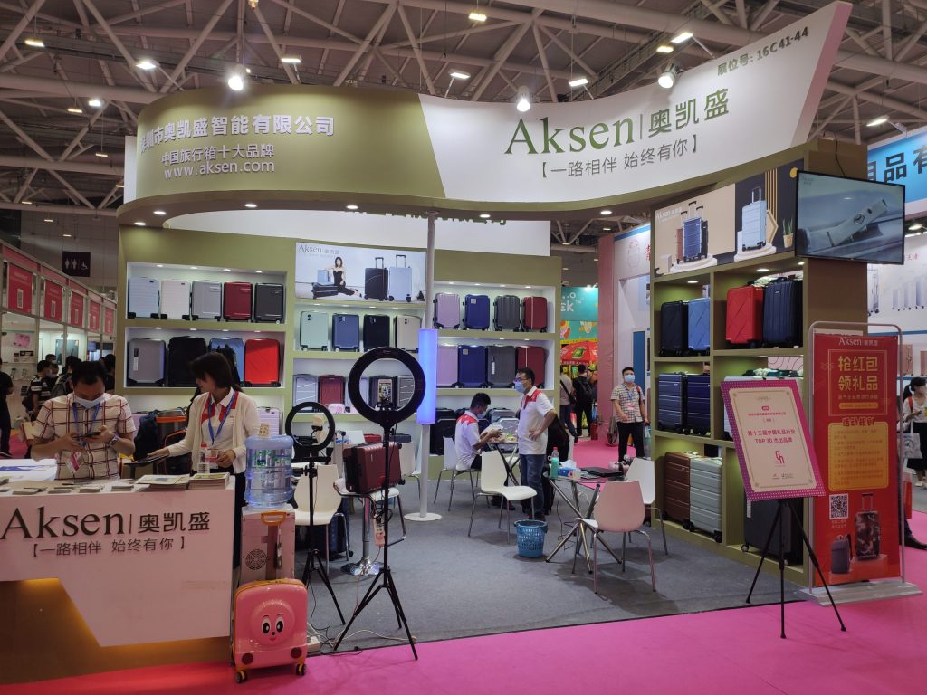 A busy trade show booth with a pink carpet, displaying many colorful suitcases. Staff interact with visitors, some wearing masks, around a counter and tables.