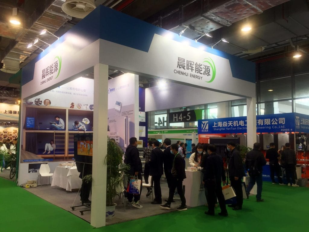 A bustling trade show booth under a white and blue canopy. Masked visitors browse displays and interact with staff around a counter, flanked by large potted plants.