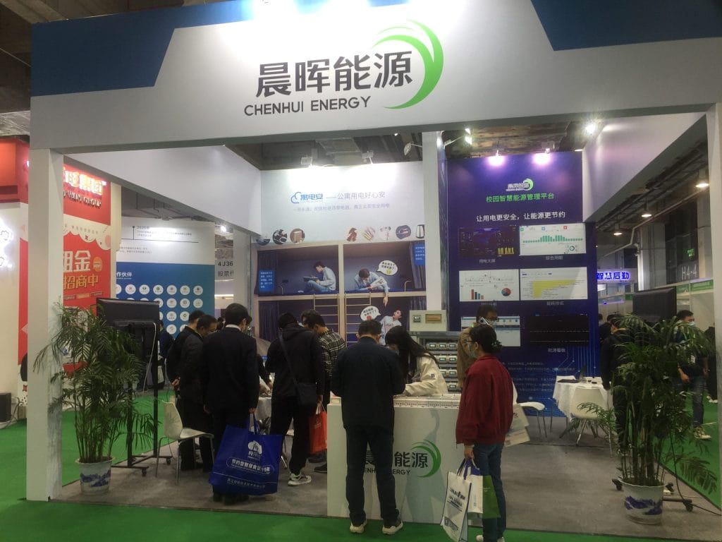 Inside a trade show booth, masked attendees gather near a reception counter, examining wall displays featuring diagrams and data charts. Potted plants are visible.