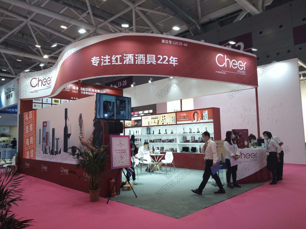 Busy red and white exhibition booth at a trade show. People walk around and interact near product displays, a screen, and a seating area on a pink floor.