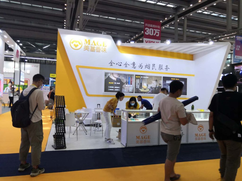 People gather at a trade show booth with white and yellow accents, product displays, and a screen showing merchandise.