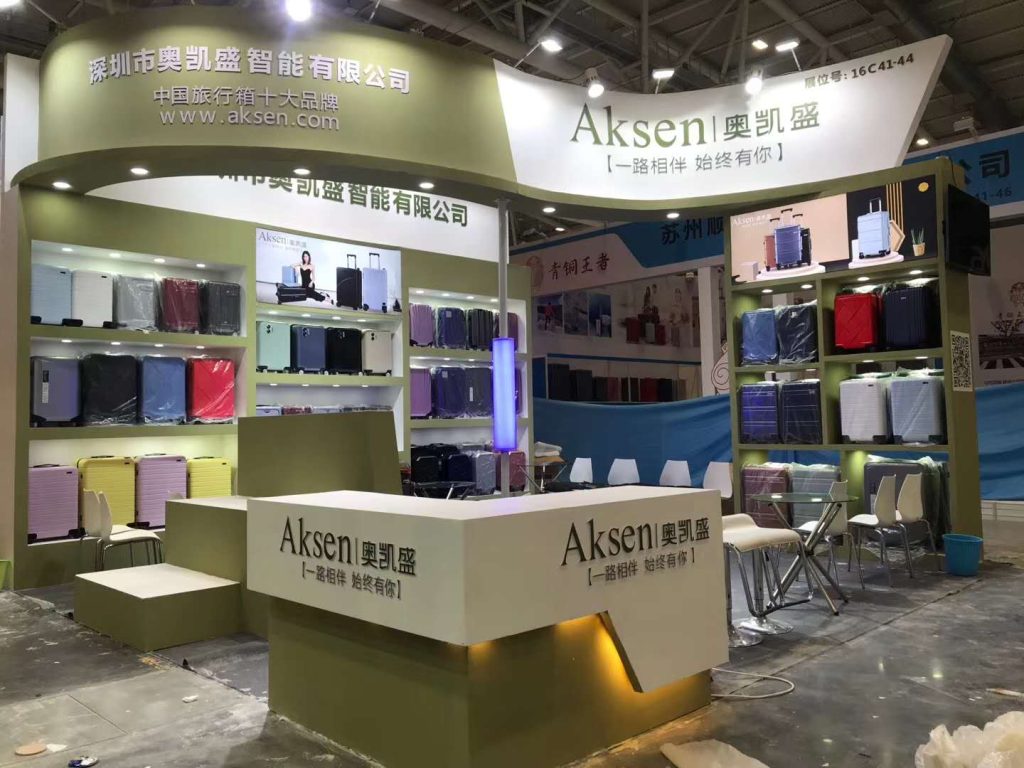 A trade show booth with green and white elements, featuring shelves filled with colorful suitcases. A white reception counter and tables with chairs are visible on a light floor.