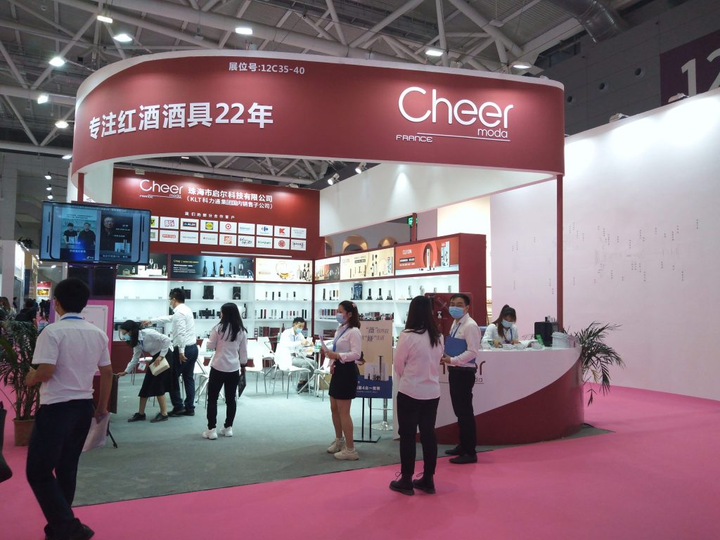 Event attendees interact inside a red and white exhibition booth with product displays, a reception desk, and a sitting area, all on a pink floor.