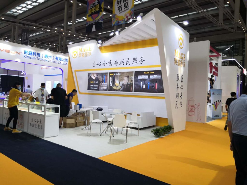 An exhibition hall features a white and yellow trade booth with product displays, a seating area, and attendees interacting.