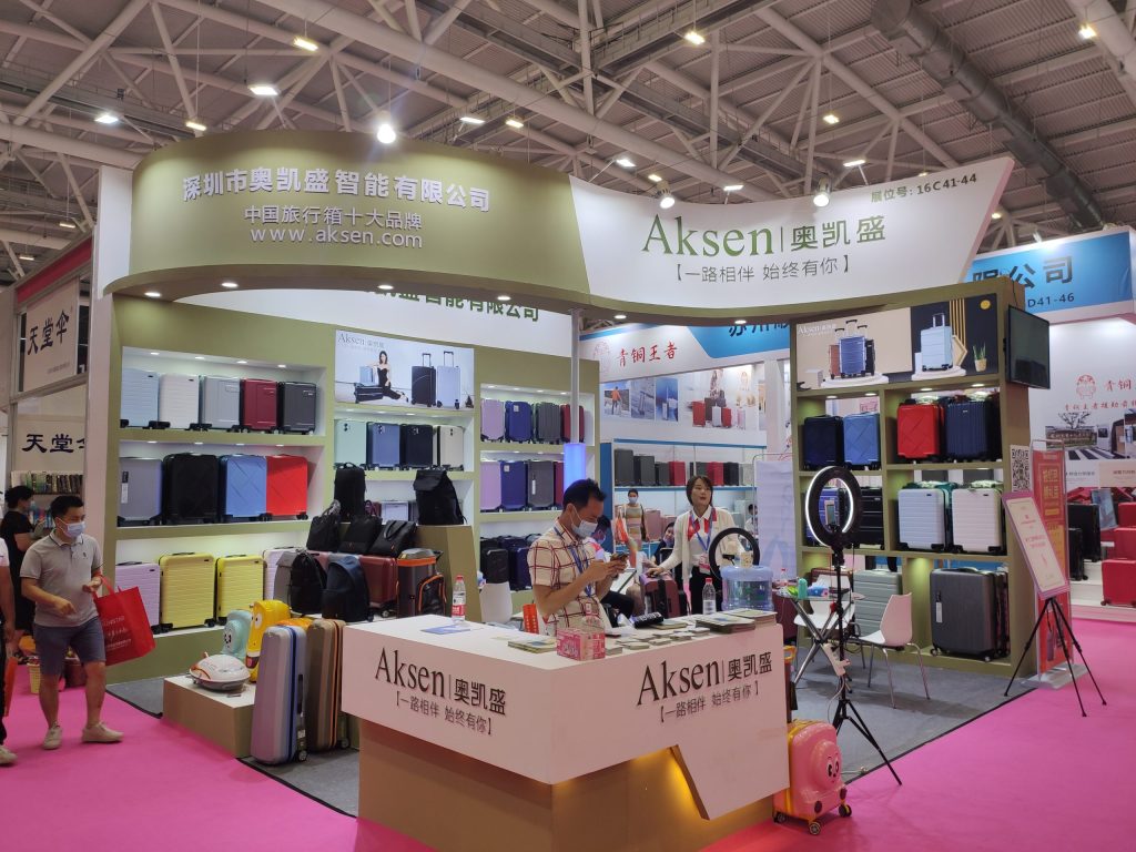 A vibrant trade show booth on a pink carpet, displaying a wide variety of colorful suitcases. Staff and visitors are present, with a ring light and promotional stands.