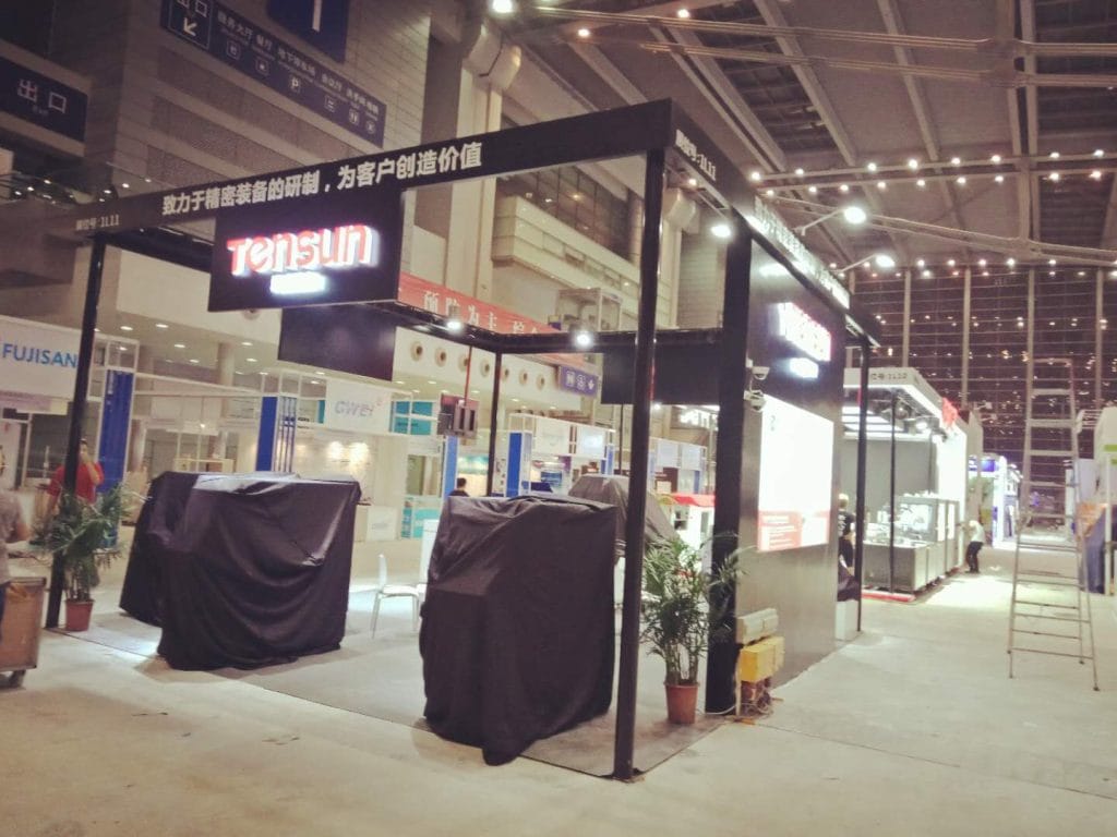 An empty exhibition booth with covered machinery, signs displaying foreign text, and plants on a concrete floor, possibly before opening.