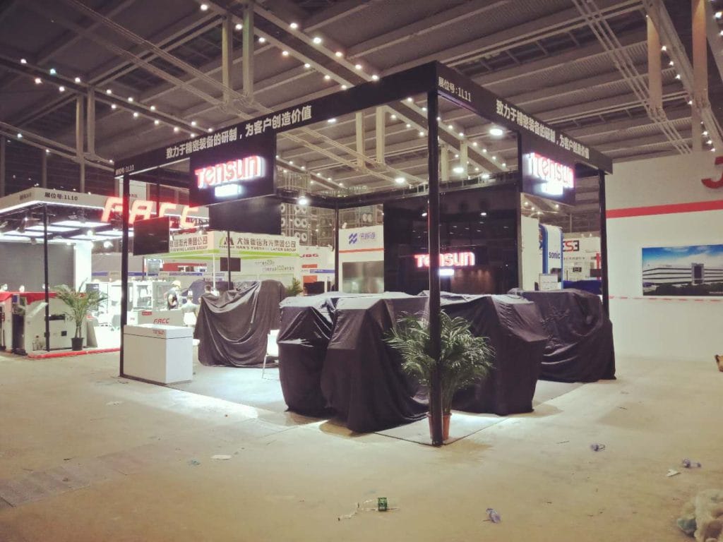 An empty exhibition booth featuring covered machinery and prominent signage, set on a concrete floor with overhead lighting.
