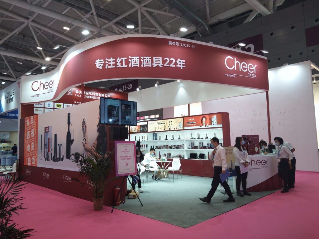 Busy red and white exhibition booth at a trade show. People walk around and interact near product displays, a screen, and a seating area on a pink floor.